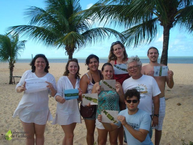 8 pessoas em pé, na praia, segurando fotos de algas, no Congresso Brasileiro de Ficologia, 2012, em João Pessoa - Paraíba. Ao fundo o céu azul claro, areia marrom claro e palmeiras.
