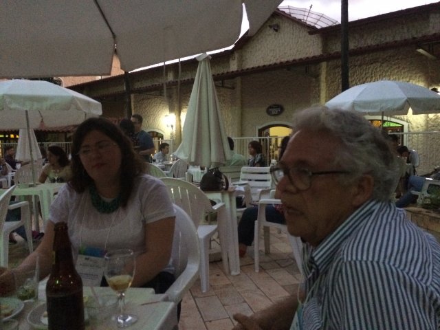 Professor Armando e Thaís sentados em uma mesa. Ao fundo outras mesas com pessoas sentadas.